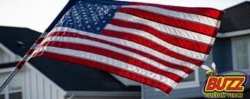 A close up of the American flag in front of houses and the logo of Buzz Custom Fence at the botto right