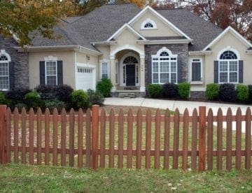 A house with trees around and a long driveway in front and brown fiberon imitation picket fence